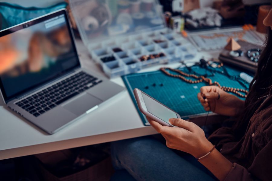 A person making beaded jewelry at a desk while looking at their smartphone.