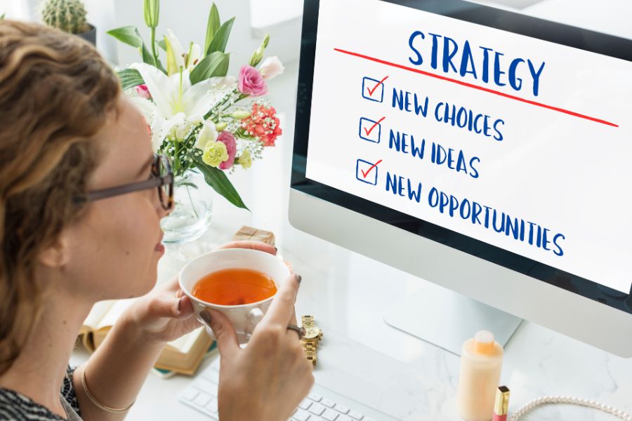 A woman at a desk drinking tea and looking at a computer screen with a "STRATEGY" checklist.