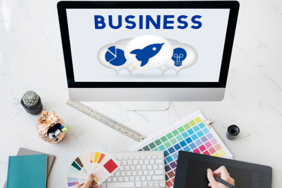 A graphic designer's desk with a computer screen that says "BUSINESS" and shows icons for a startup.