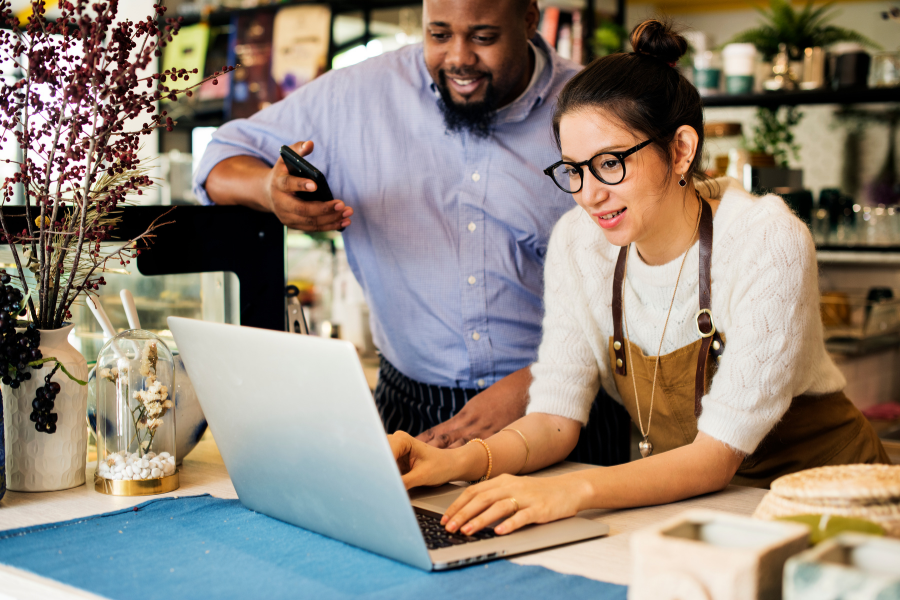 A man and woman collaborate on a laptop at a coffee shop, surrounded by warm lighting and cozy seating.