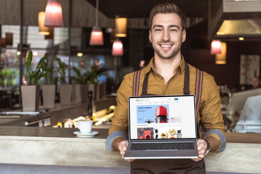 A smiling cafe owner in an apron holds up a laptop showing the website homepage.