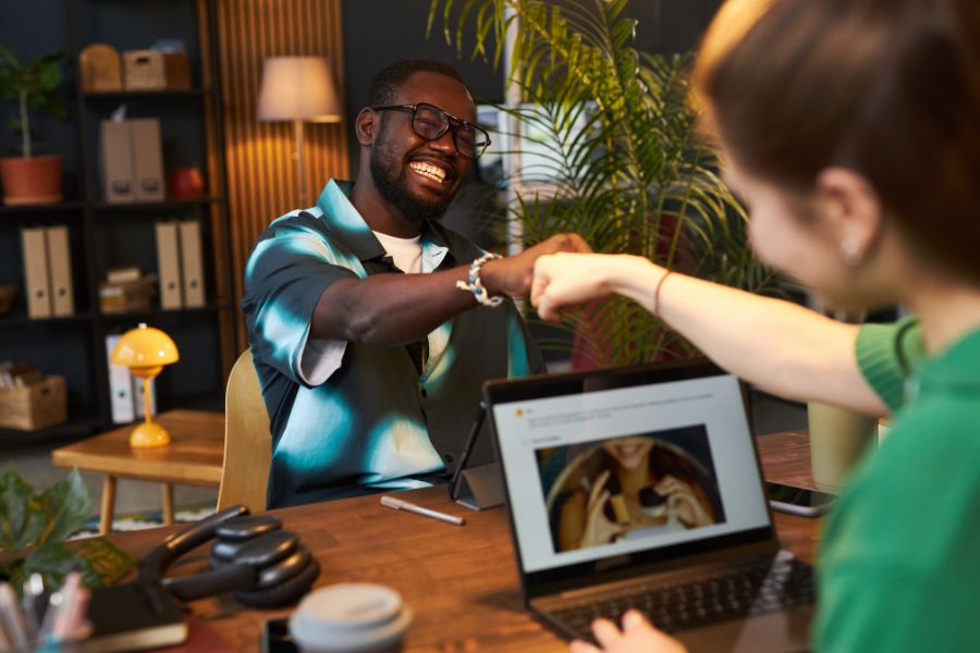 Two colleagues give each other a fist bump over a desk in an office.